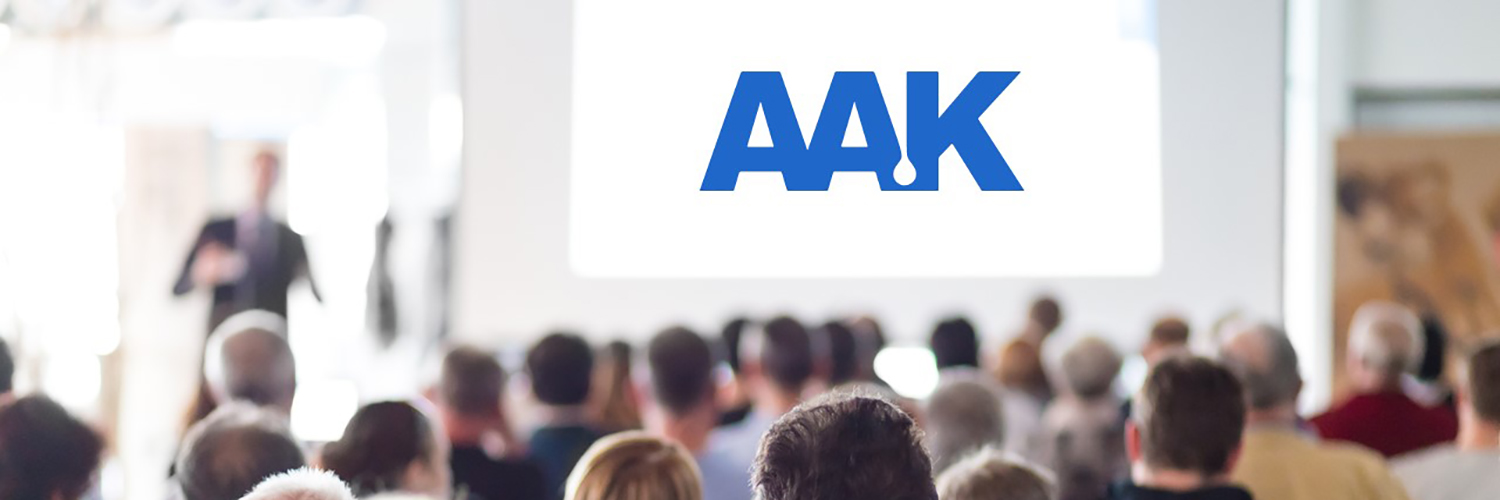 Group seated facing large screen displaying 'AAK' in blue, with a person standing near the screen giving a presentation.