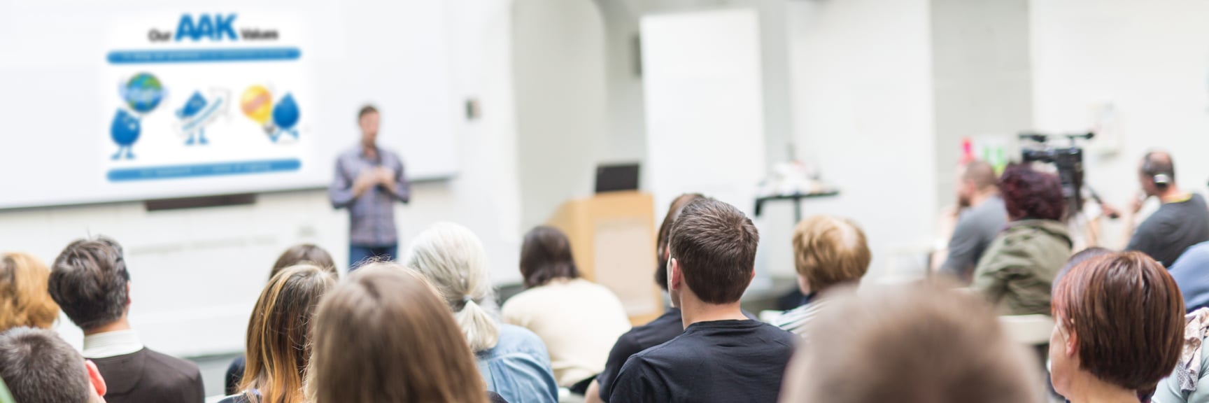 People seated facing a presenter in a well-lit seminar room. The screen displays “Our AAK Values”.