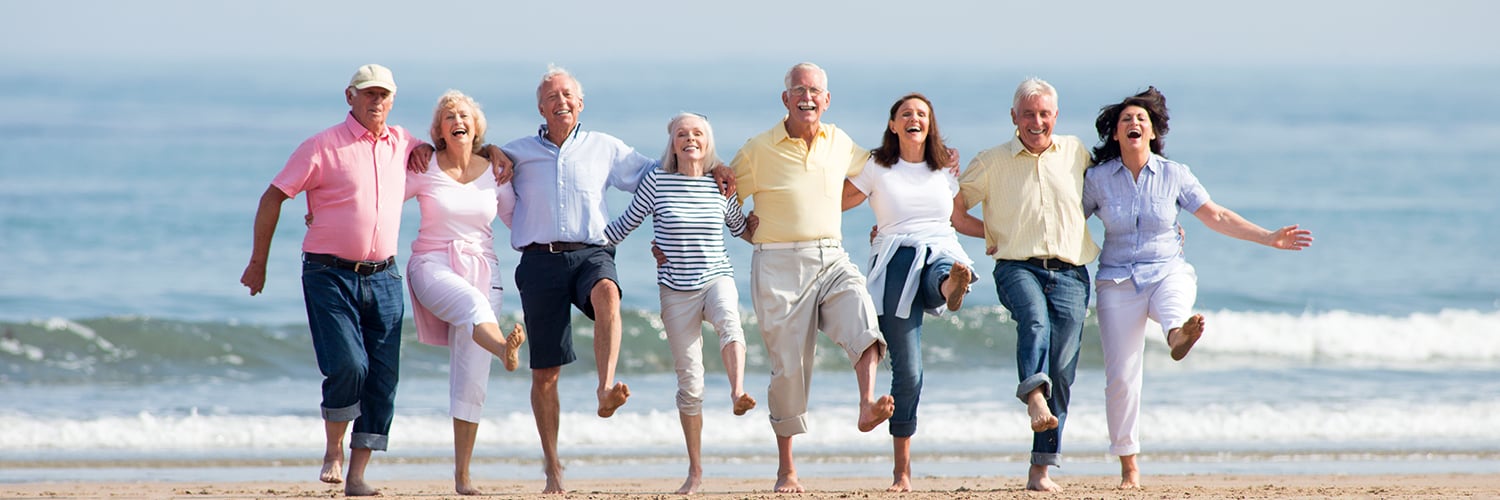 Eight seniors stand in a row on the beach, smiling, holding each other, lifting one leg.