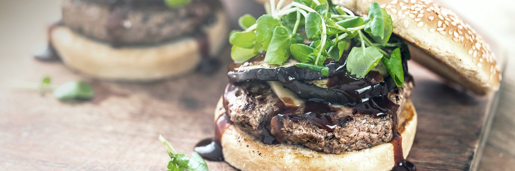 Close-up of a plant-based meat burger with greens, grilled vegetables, and sauce on a sesame bun, placed on a wooden surface.