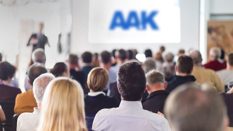 Grupp sitter vänd mot stor skärm med 'AAK' i blått, en person står vid skärmen och håller en presentation.