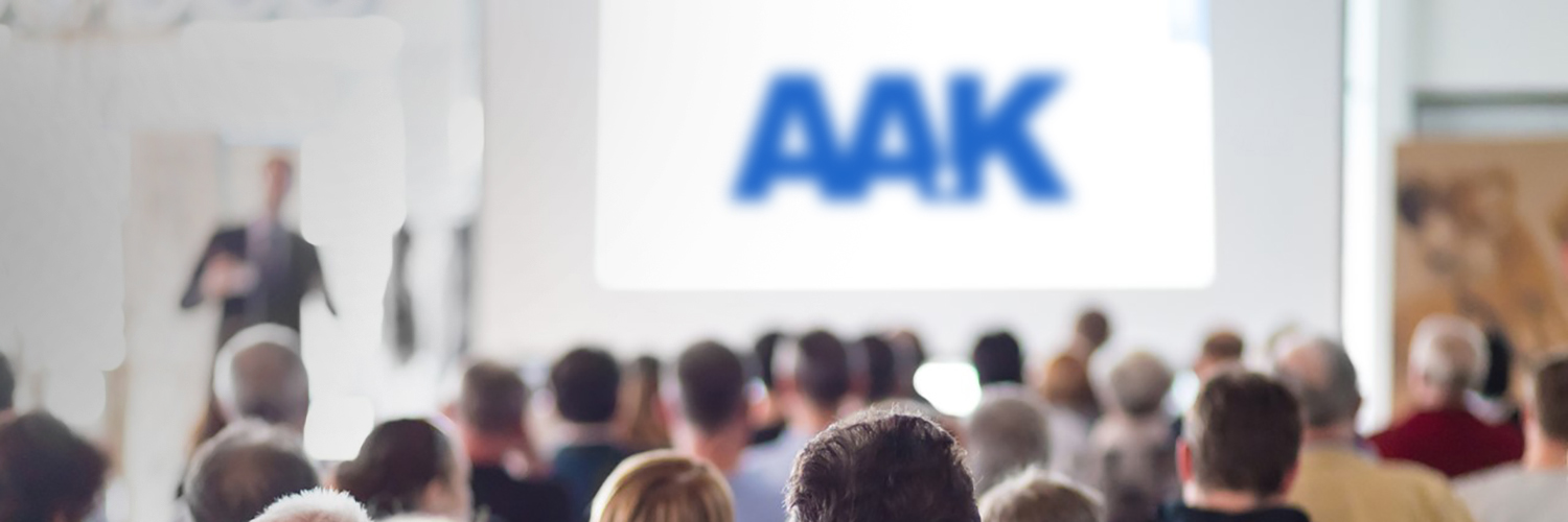 Group seated facing large screen displaying 'AAK' in blue, with a person standing near the screen giving a presentation.