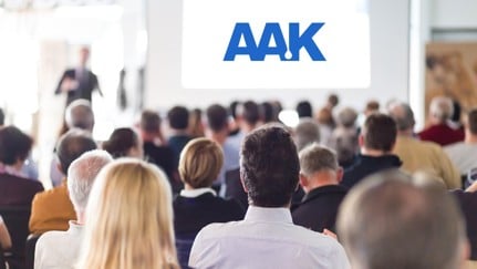 Group seated facing large screen displaying 'AAK' in blue, with a person standing near the screen giving a presentation.