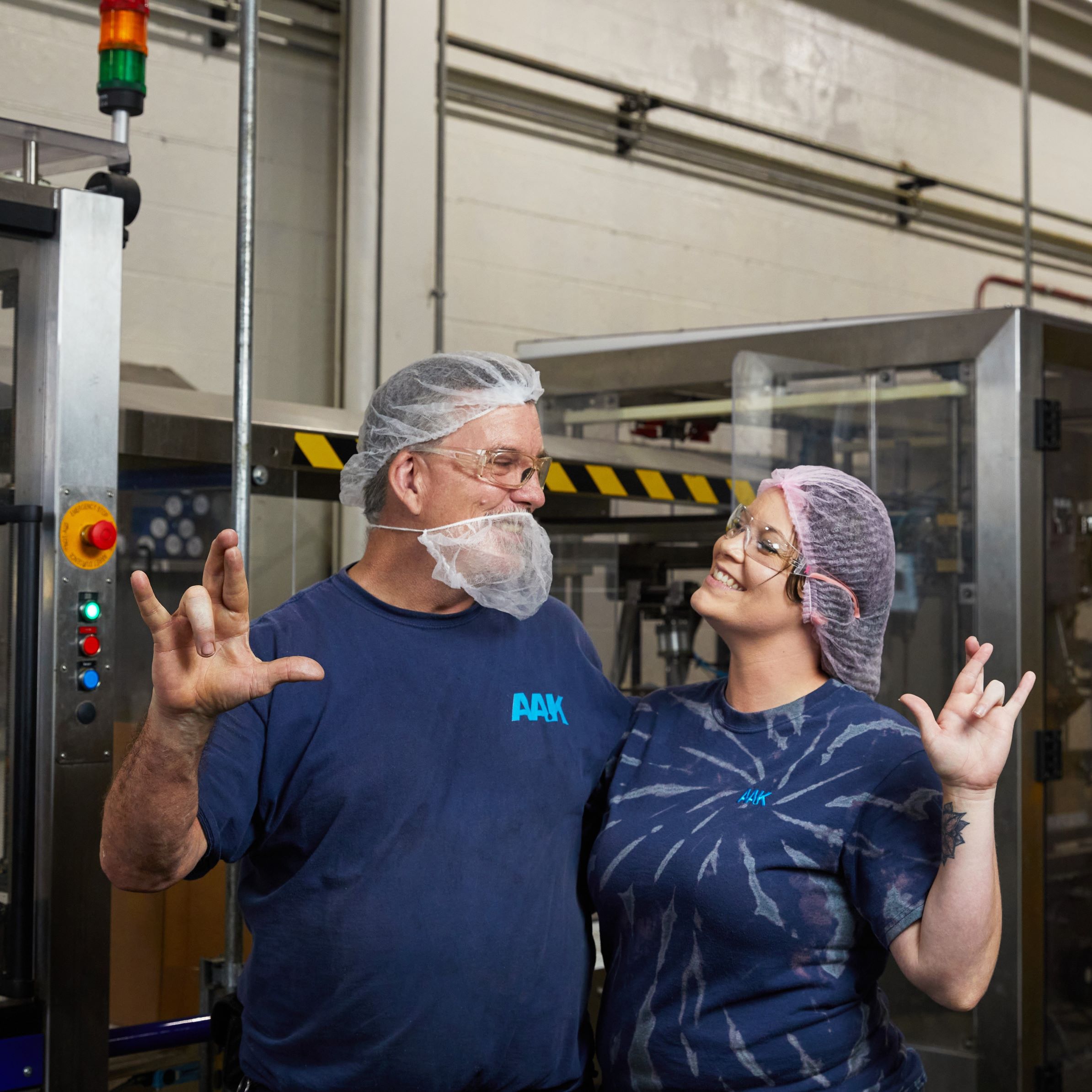 Two AAK employees wearing blue shirts and hairnets, standing in an industrial setting and making hand gestures.