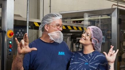 Two AAK employees wearing blue shirts and hairnets, standing in an industrial setting and making hand gestures.