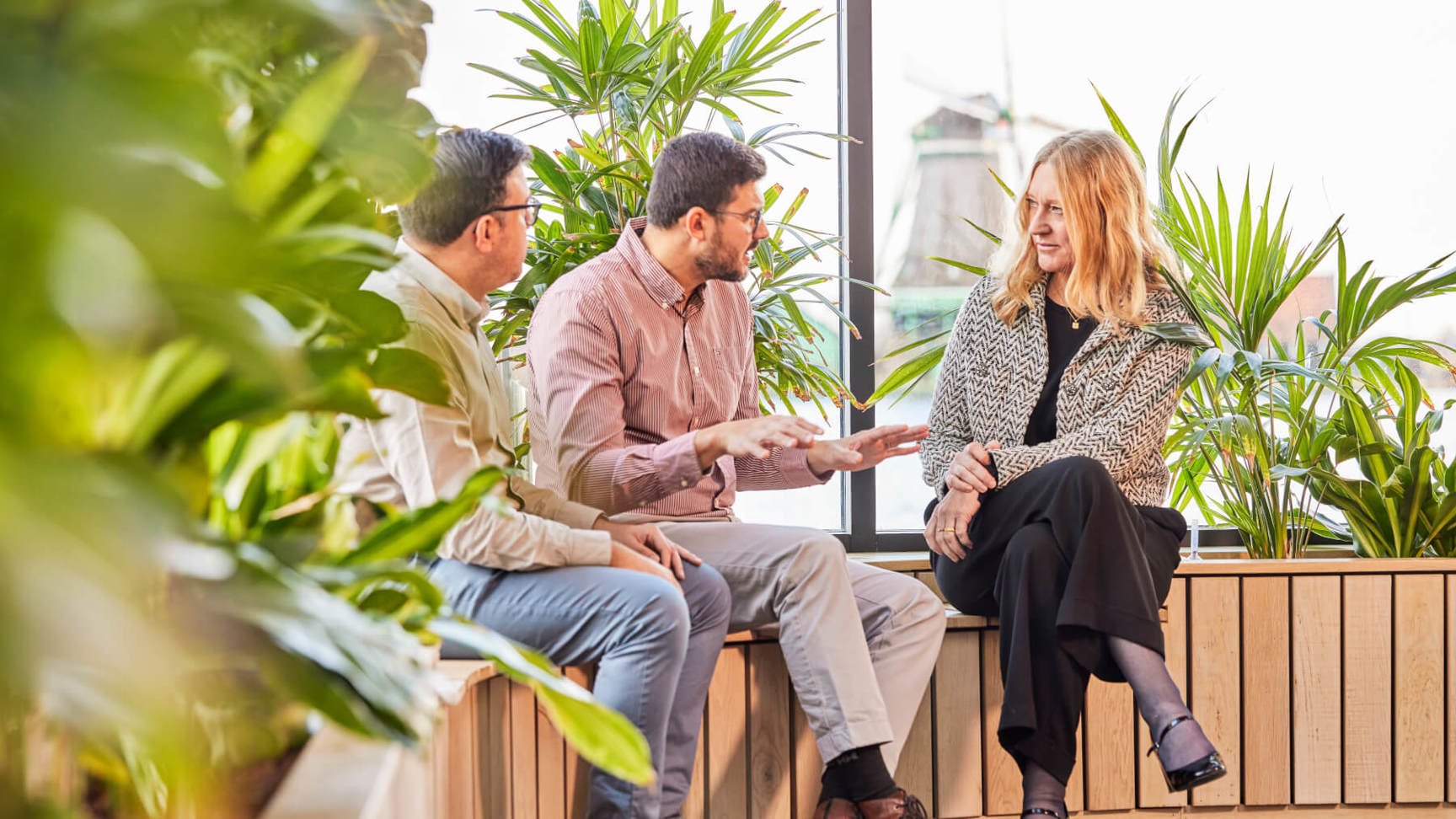 hree people engaged in conversation while sitting on a bench inside an office environment.
