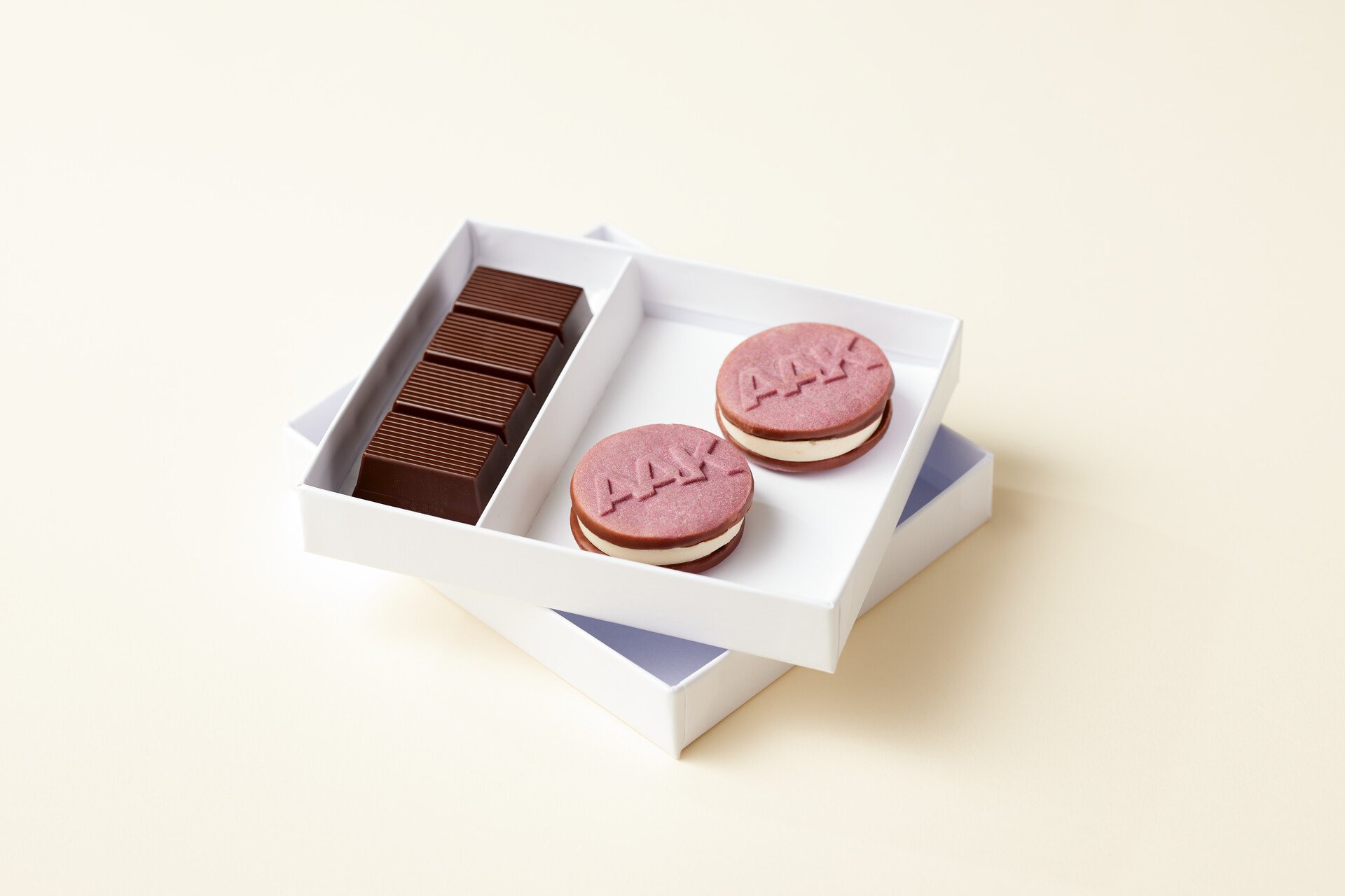 Open white box with two raspberry-colored sandwich cookies embossed with “AAK” and a row of rectangular chocolate pieces
