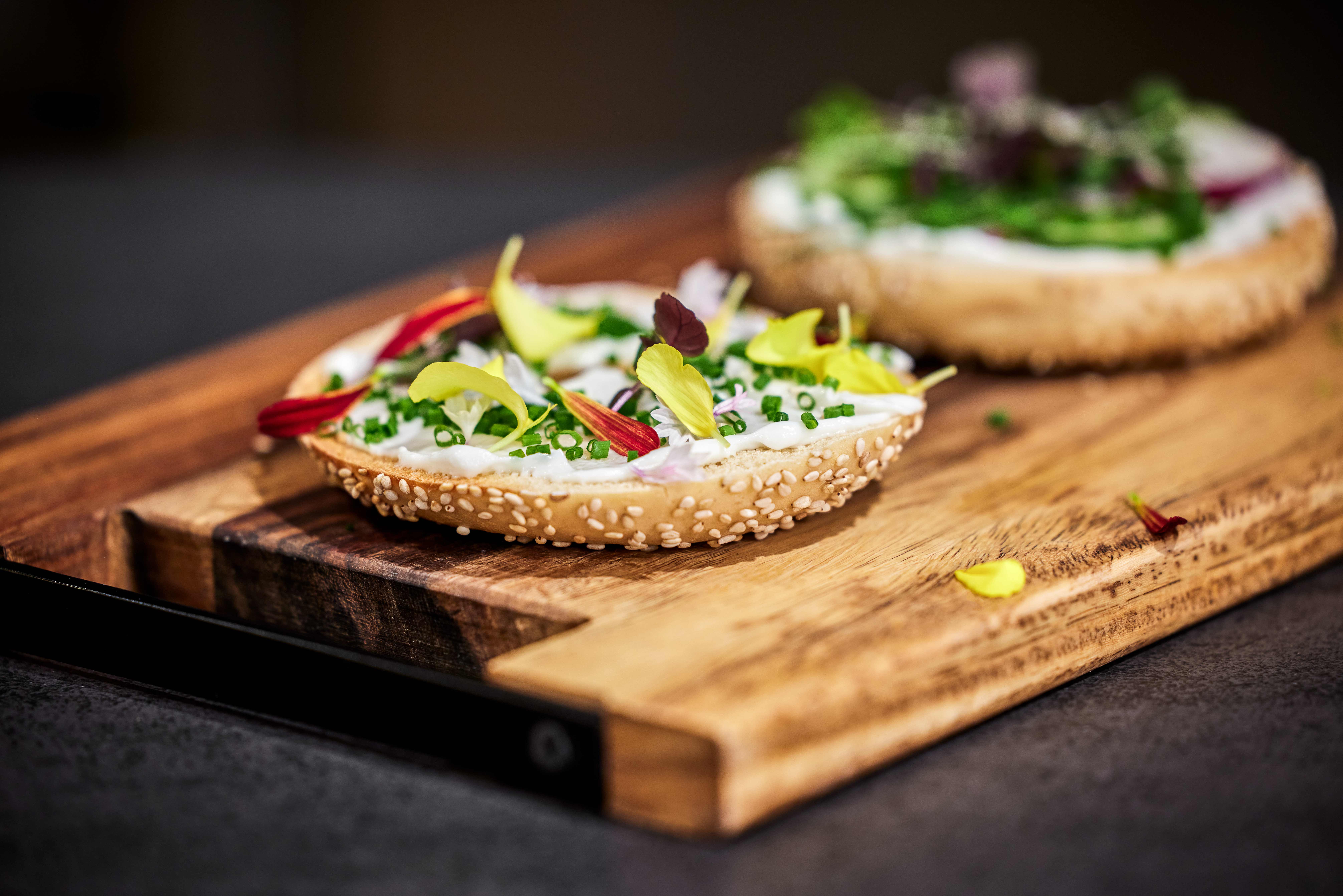 Close-up of a sesame bagel topped with plant based cream cheese, fresh herbs, and colorful edible flowers on a wooden board