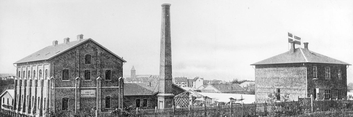 Svartvit bild av Århus Palmkärnefabrik, AAK:s första industriplats med tegelbyggnader och hög skorsten i stadsmiljö.