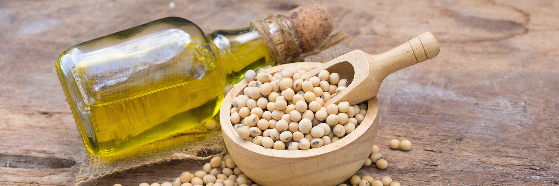 Wooden bowl of soybeans with scoop beside a glass bottle of soybean oil on a rustic wooden surface.