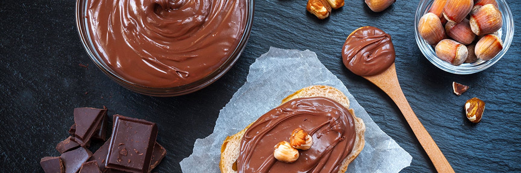 Chocolate hazelnut spread on bread, with hazelnuts, dark chocolate, and a wooden spoon.