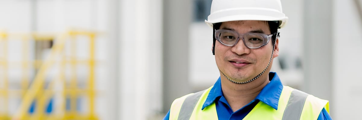 Alex Wang, HSE Manager in China, wearing a white hard hat and yellow safety vest in an industrial setting with yellow structures.