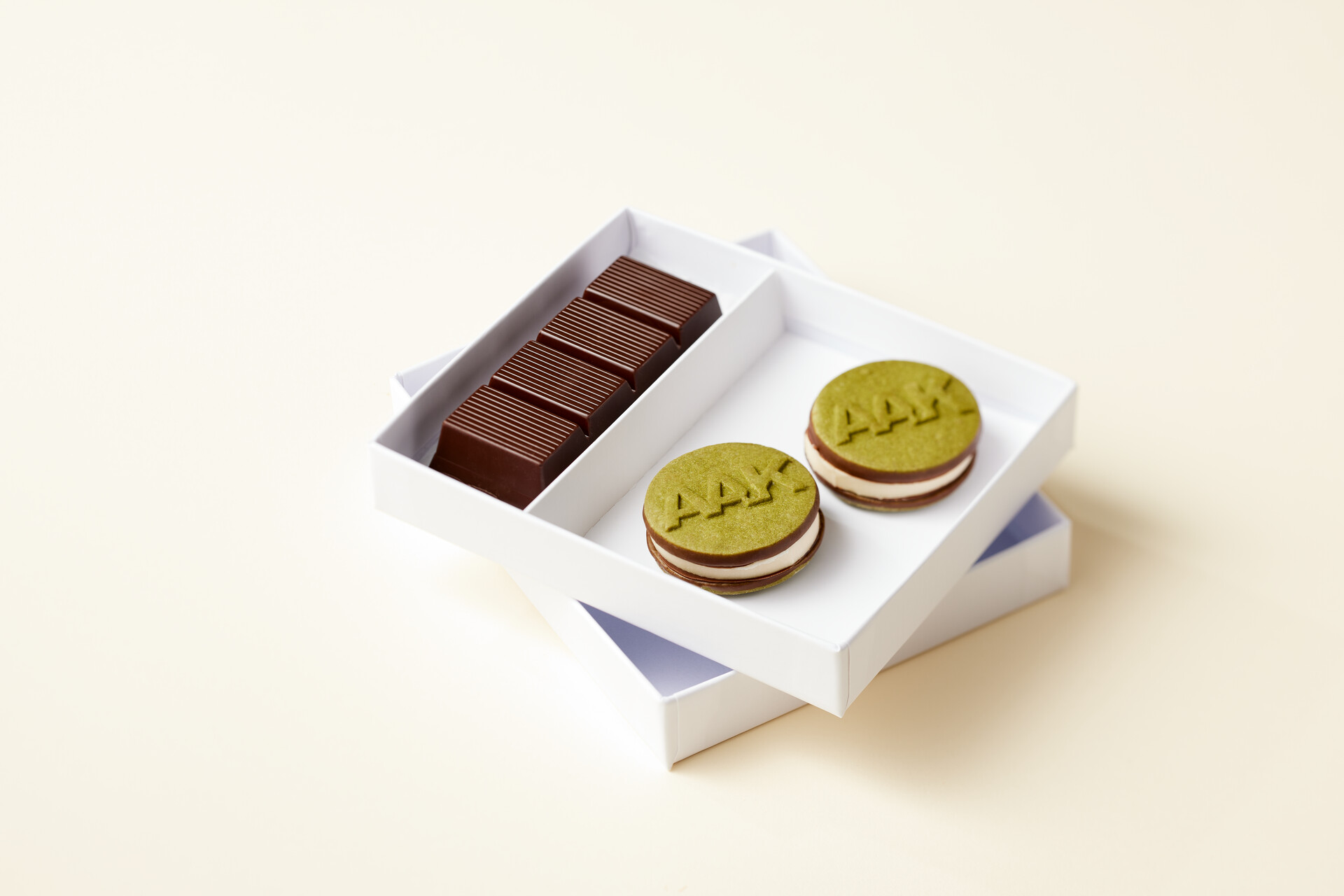 Open white box with two pistachio-green sandwich cookies embossed with “AAK” and a row of rectangular chocolate pieces