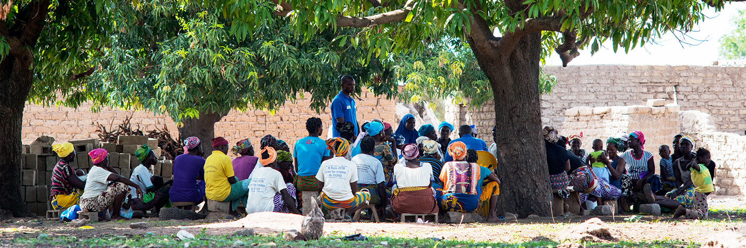 Group of people in Kolo Nafaso sitting in a circle under trees, engaged in discussion; many wear colorful clothing and headscarves.