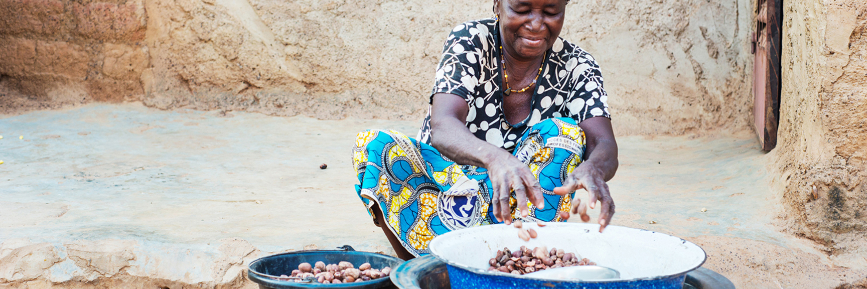West african woman sitting on the ground sorting shea kernels in large bowls, smiling.