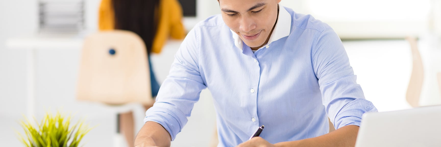 Man in a blue shirt sitting at a desk writing with a pen, with a laptop and a green plant in front of him in a bright office space.