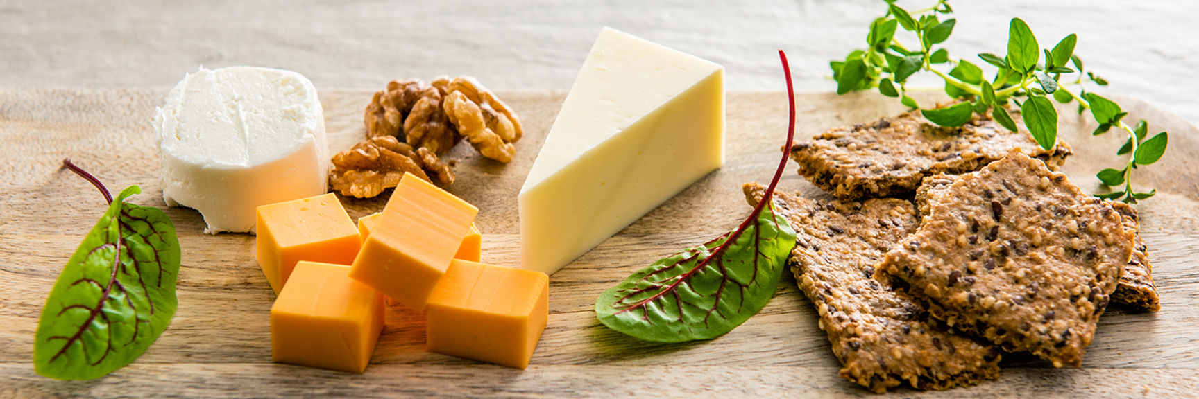 Cheese board with soft and hard cheeses, walnuts, crackers, herbs, and leafy greens.