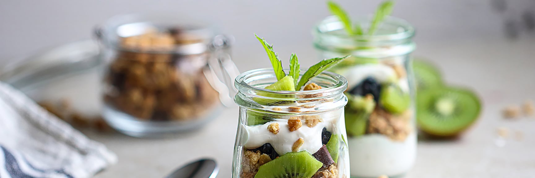 Three glass jars with layered plant-based yogurt, granola, kiwi, and blueberries, topped with mint; spoon and napkin in foreground.