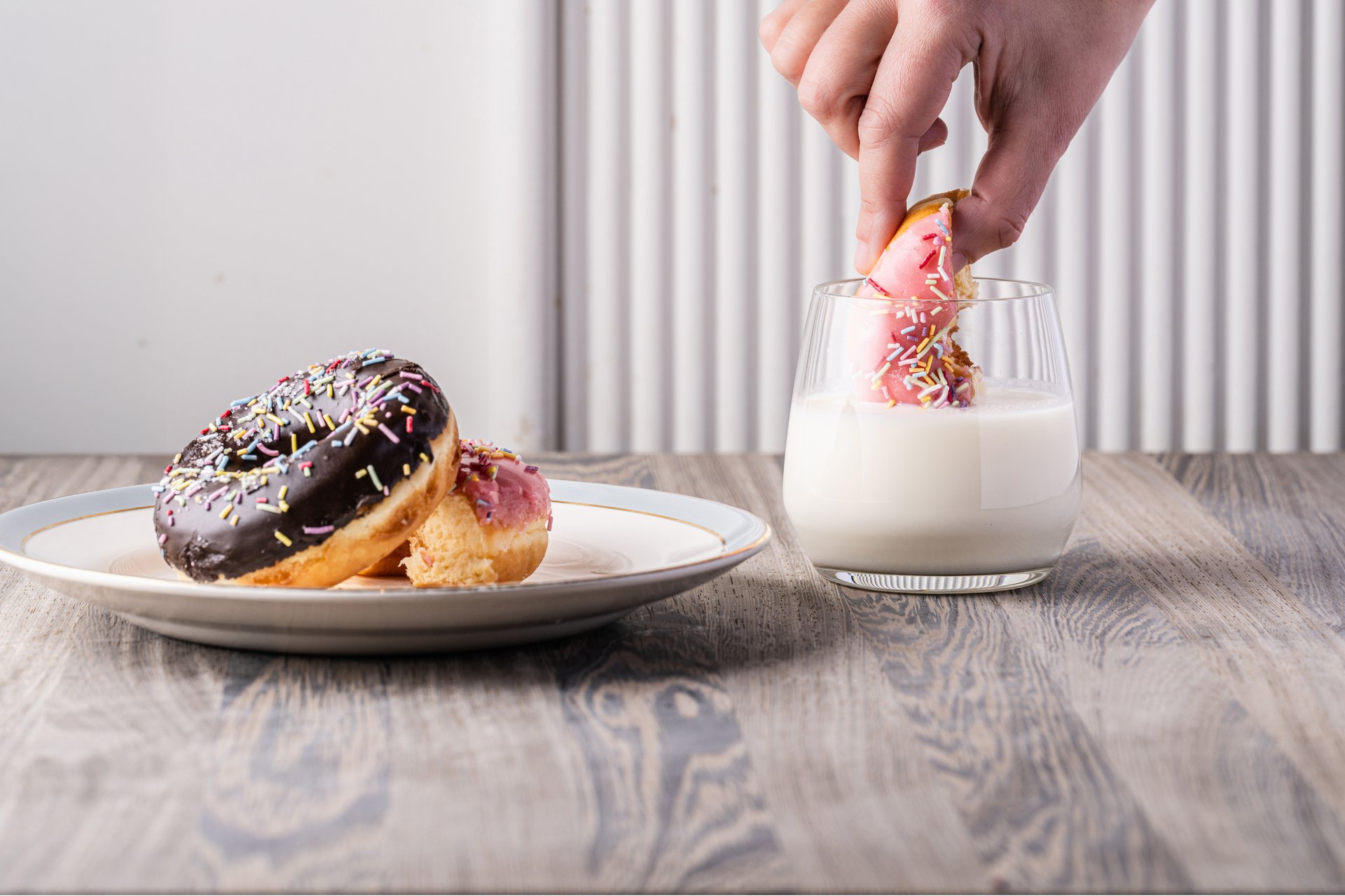 A hand dipping colorful, sprinkle-covered donuts into creamy plant-based milk
