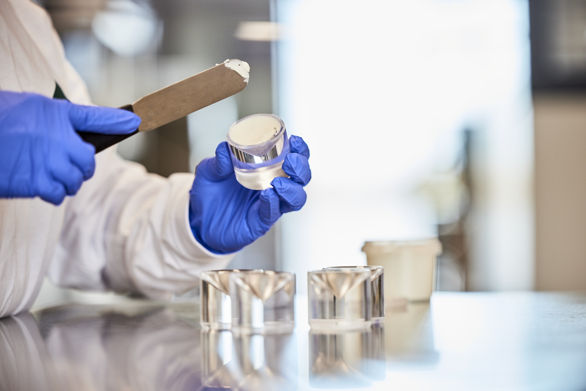 Person wearing blue gloves applying a creamy substance into a small transparent container in a lab setting