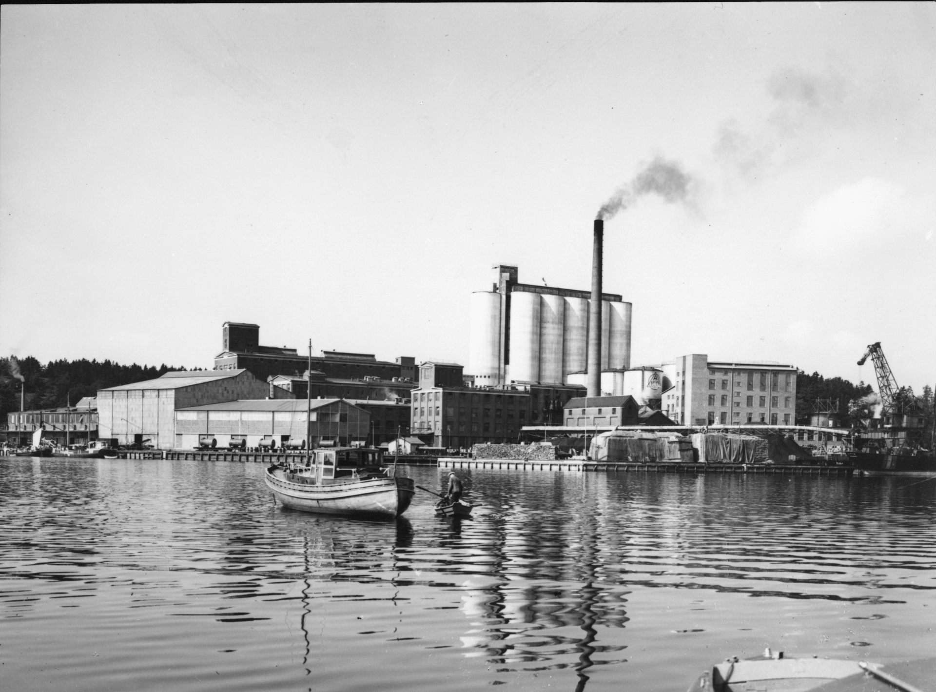 Black and white photo of the factory in Karlshamn by the waterfront, with large silos, a tall chimney, and boats in the foreground