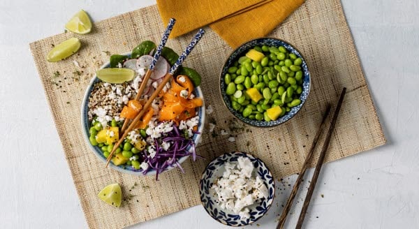 A bowl filled with plant based dairy feta cheese, vegetables and chopsticks resting on a woven mat.