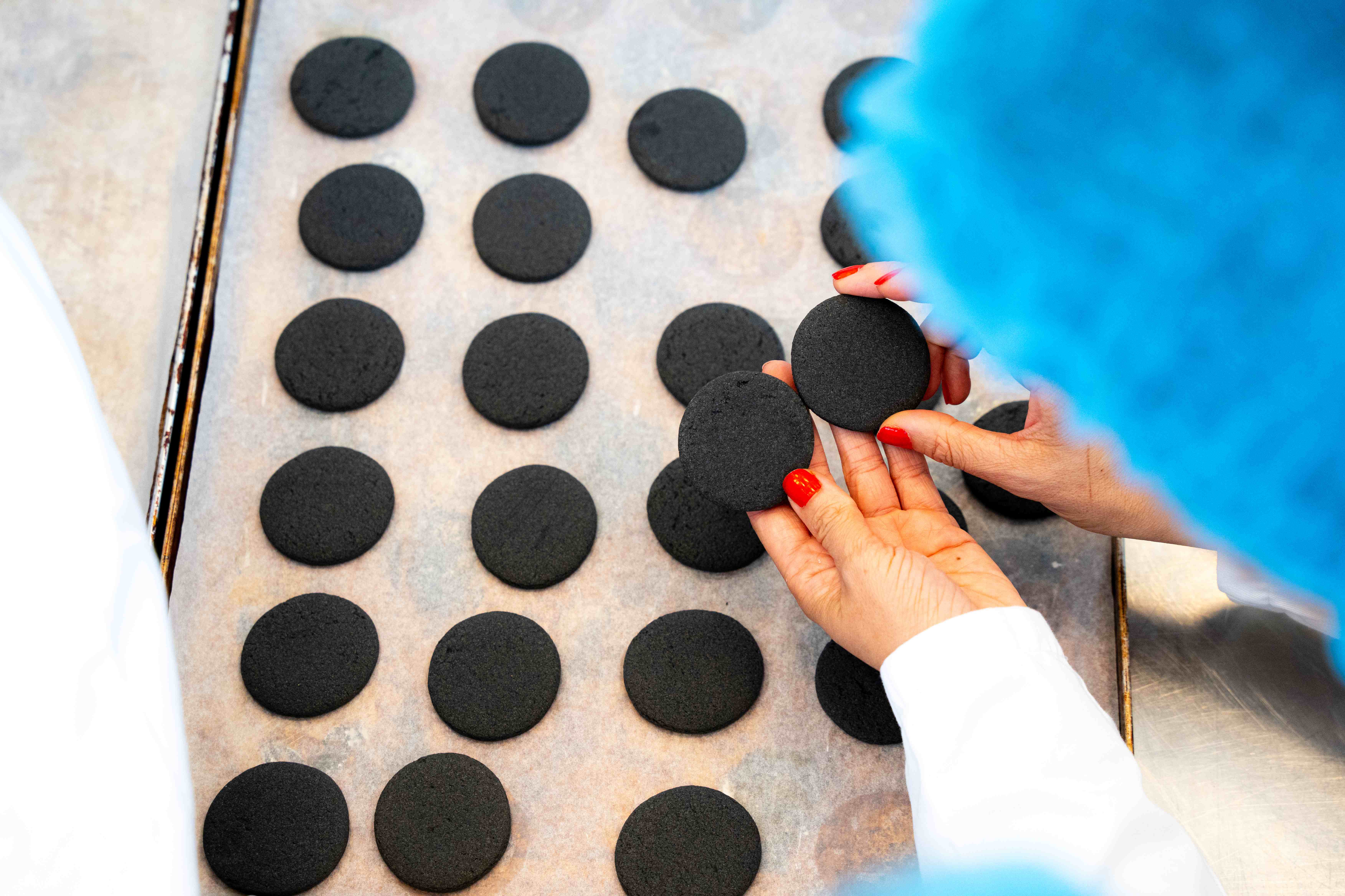Person in lab clothing holding and inspecting dark round cookies on a baking tray.