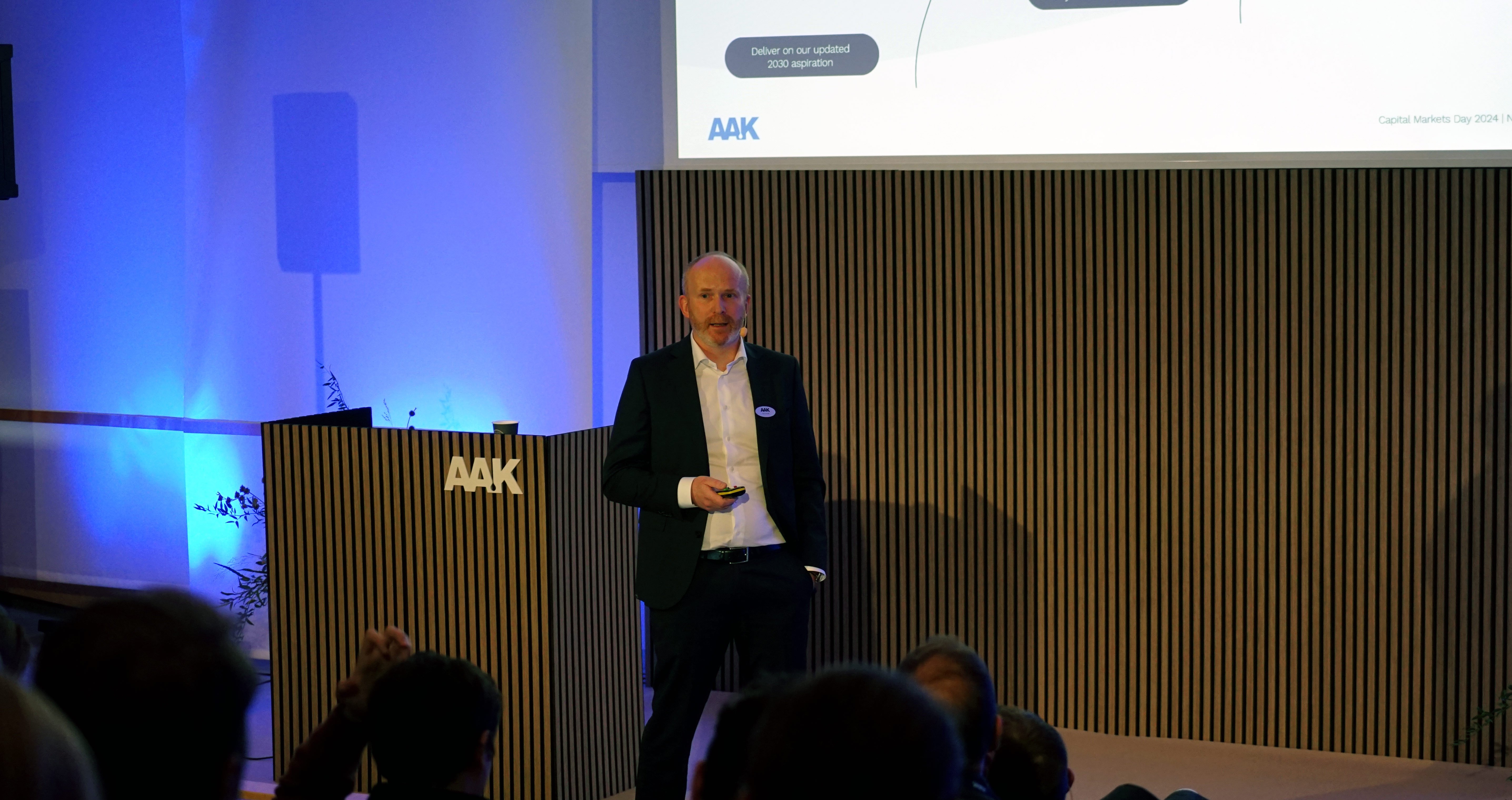 Man on stage presenting with AAK logo on screen and podium, audience seated in front.