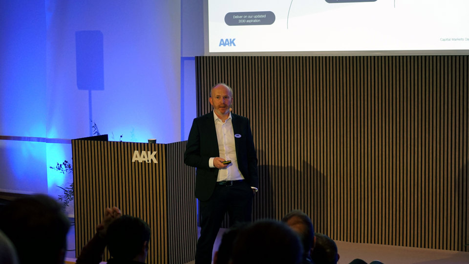 Man på scen presenterar med AAK-logga på skärm och podium, publik sitter framför.