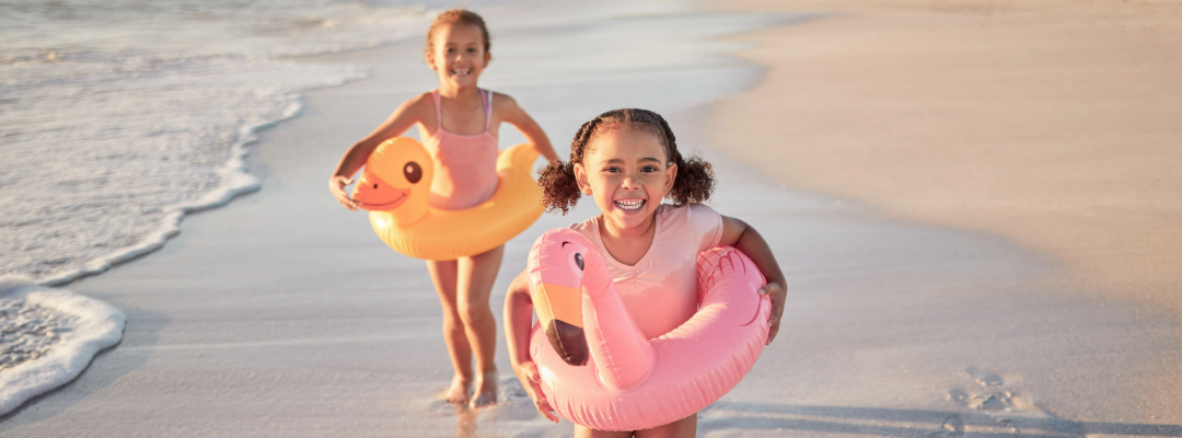 Two children on a beach holding inflatable toys a pink flamingo and a yellow duck with ocean waves in the background.