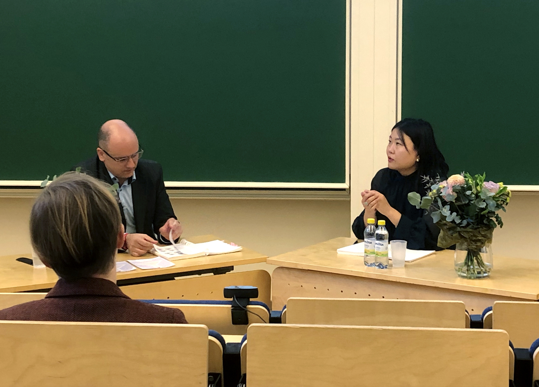 Two people seated at a desk in a lecture hall with a green chalkboard behind.