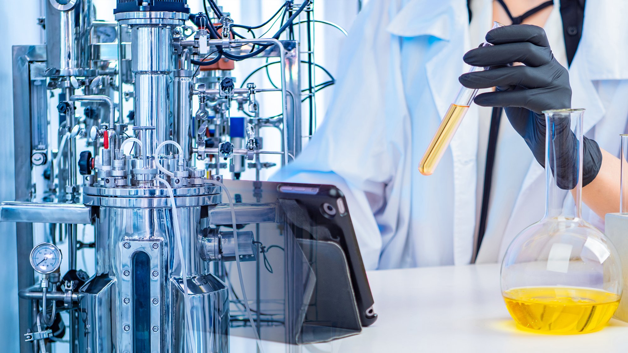 A person in a lab coat holds a test tube of oil in a laboratory with equipment.